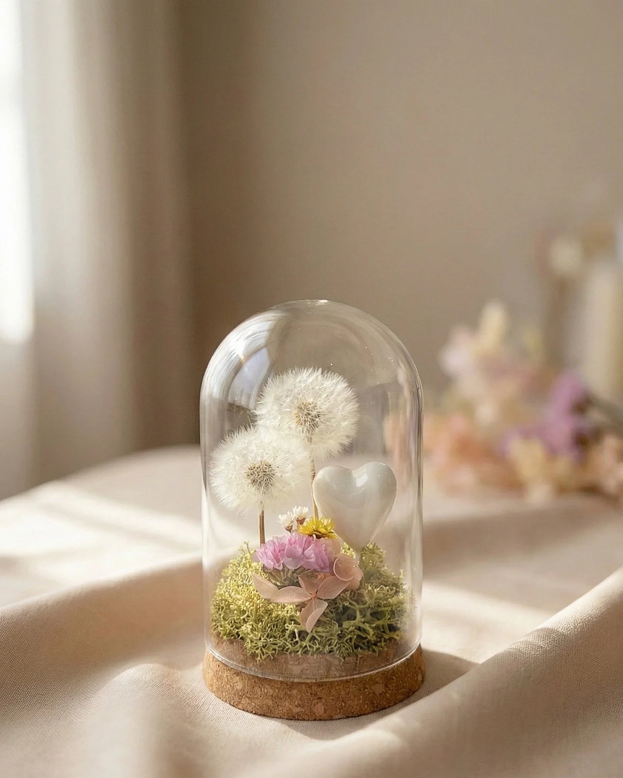 composizione con due dente di leone ( soffioni ) preservati in campana di vetro con base di sughero con glixie, ortensia e statice color pastello e cuoricino bianco e muschio verde stabilizzato